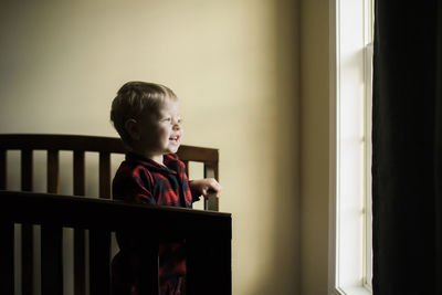 Smiling baby boy looking through window while standing in crib at home