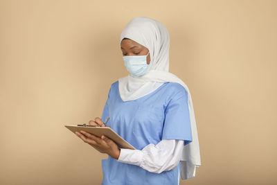 A young muslim nurse standing in a surgical mask takes note on a clipboard on a yellow wall