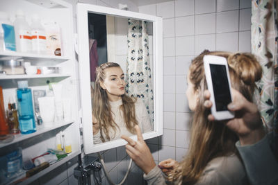 Cropped hand of woman photographing female friend in mirror reflection at dorm bathroom