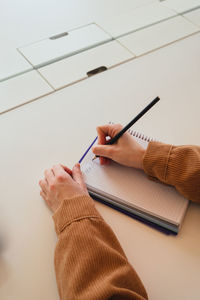 From above cropped unrecognizable woman in casual clothes with hair bun touching head and writing in planner while sitting at table and studying in university library