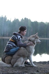 Side view of young woman sitting with siberian husky at lakeshore