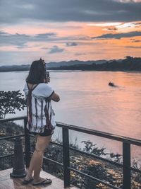 Woman standing on railing against sky during sunset