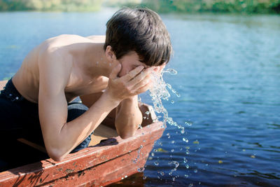 Shirtless boy washing face from lake while sailing on boat