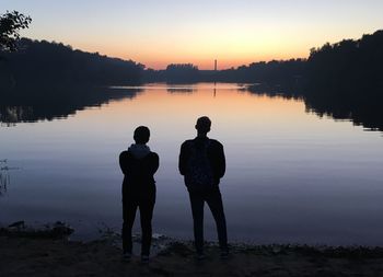 Rear view of silhouette men standing by lake against sky during sunset