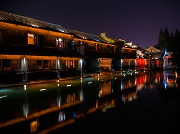 Illuminated buildings by lake against sky at night