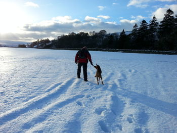 Full length rear view of man with dog walking on snowy landscape during sunny day