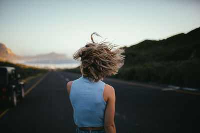 Rear view of woman with tousled hair standing on road against clear sky