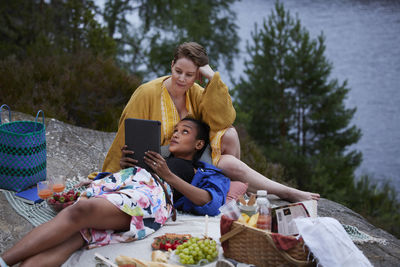 Female couple having picnic by river and using tablet