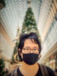 Portrait of young man in eyeglasses and face mask against christmas tree and skylight inside mall.