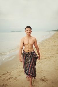 Portrait of shirtless man standing at beach against sky