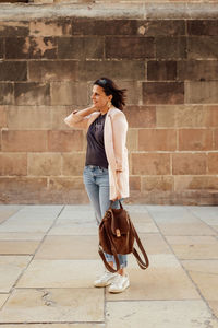 Full body of positive adult female in trendy casual outfit with backpack in hand standing against aged stone wall on urban street