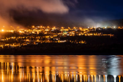 Illuminated buildings by lake against sky at night