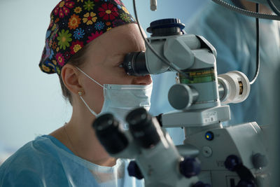 Focused adult female doctor in sterile mask and ornamental medical cap looking through surgical microscope against crop coworker in hospital