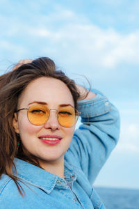 Portrait of young diverse woman in yellow glasses smiling, standing by seaside. generation z.