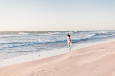 Young woman walking at shore against clear sky during sunset