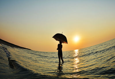 Silhouette woman with umbrella standing in sea against orange sky