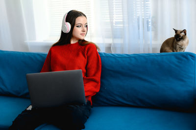 A young girl in a red  sweater works on a laptop at home. her cat and dog are sitting next to her