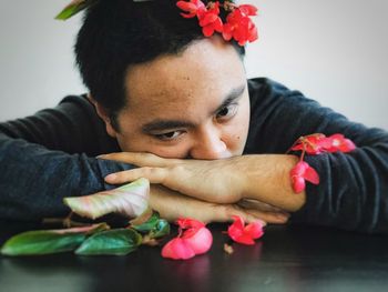 Close-up of young man with red begonia flowers resting his head on forearms on table.