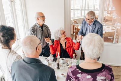 Senior men holding hands with senior woman while friends standing by table in restaurant