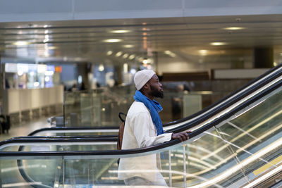 African american passenger man with suitcase stands on escalator, holds handrail in airport terminal