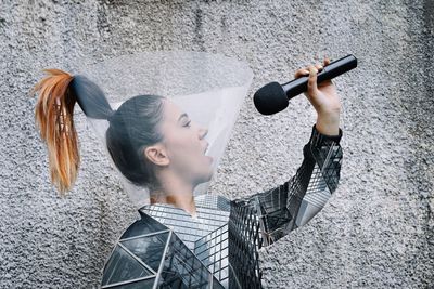 Young woman with mask singing and holding microphone in hands against wall