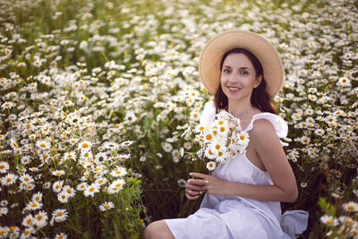 Beautiful young brunette woman in a hat and a white dress standing on a chamomile field at sunset