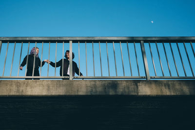 Low angle view of friends talking while standing by railing on bridge against clear sky