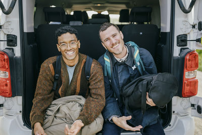 Portrait of happy male friends sitting with luggage in van trunk