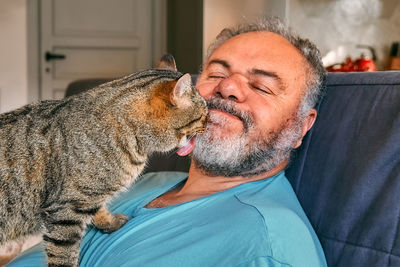 Tabby cat licking face of bearded man in living room. human-animal relationships. pets care.