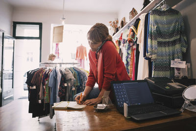 Woman writing in diary while talking on smart phone at store
