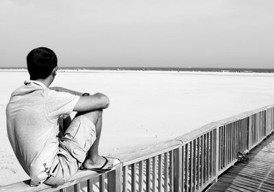 Rear view of man sitting on railing at beach against sky