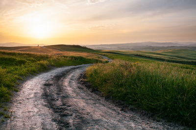 Dirt road amidst field against sky during sunset