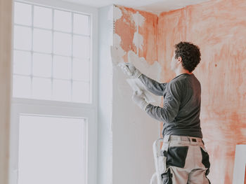 A young caucasian guy applies fresh putty with a small spatula on the wall near the window