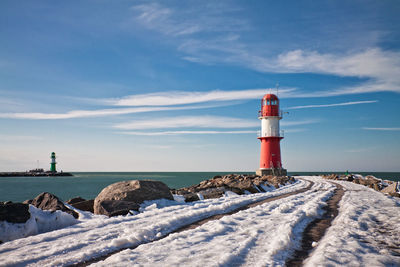 Snow covered pier towards lighthouse at sea against blue sky