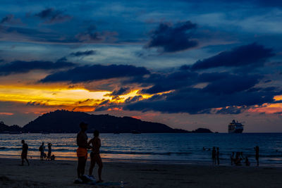 Silhouette people on beach against dramatic sky during sunset