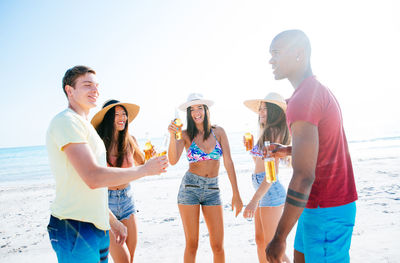 Friends holding drinks while standing at beach against sky