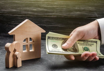 The hand of a businessman stretches a wad of money to the family figures near the wooden house. 