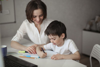 Beautiful woman tutor is engaged with a child boy at home at a table, real interior