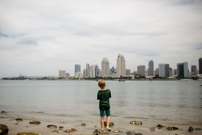 Rear view of boy against sea and cityscape against sky