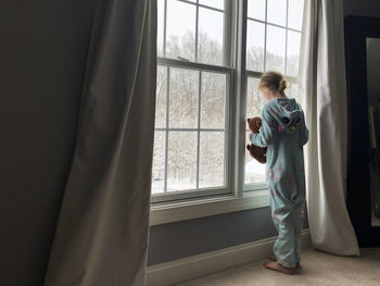 Rear view of girl in pajama onesie holding teddy looking out window