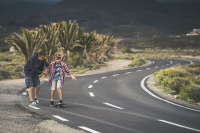 Man assisting son skateboarding on road against mountains