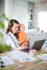 Busy female parent running business while taking daughter's care at dining table in kitchen