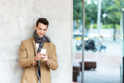 Young man using smart phone while standing against wall