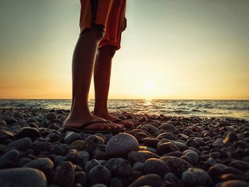 Low section of person standing on pebbles at beach against sky during sunset