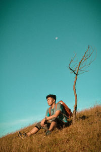 Young man sitting on field against clear blue sky