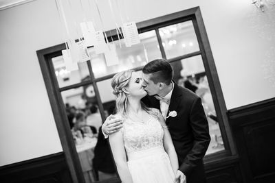 Newlywed couple kissing against window