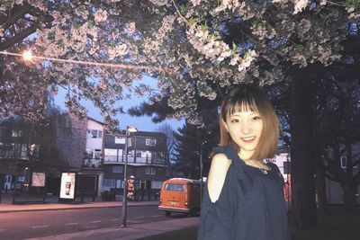 Portrait of smiling woman standing against tree on city street at night