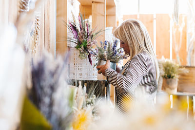 Side view of blond senior woman in striped blouse enjoying smell of fresh flowers in metal pot while buying plants in store