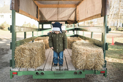 Full length of boy standing on vehicle trailer by hay bales at farm