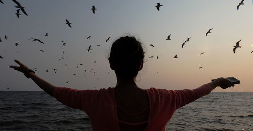 Rear view of woman with arms outstretched looking at birds flying over seascape against sky during sunset